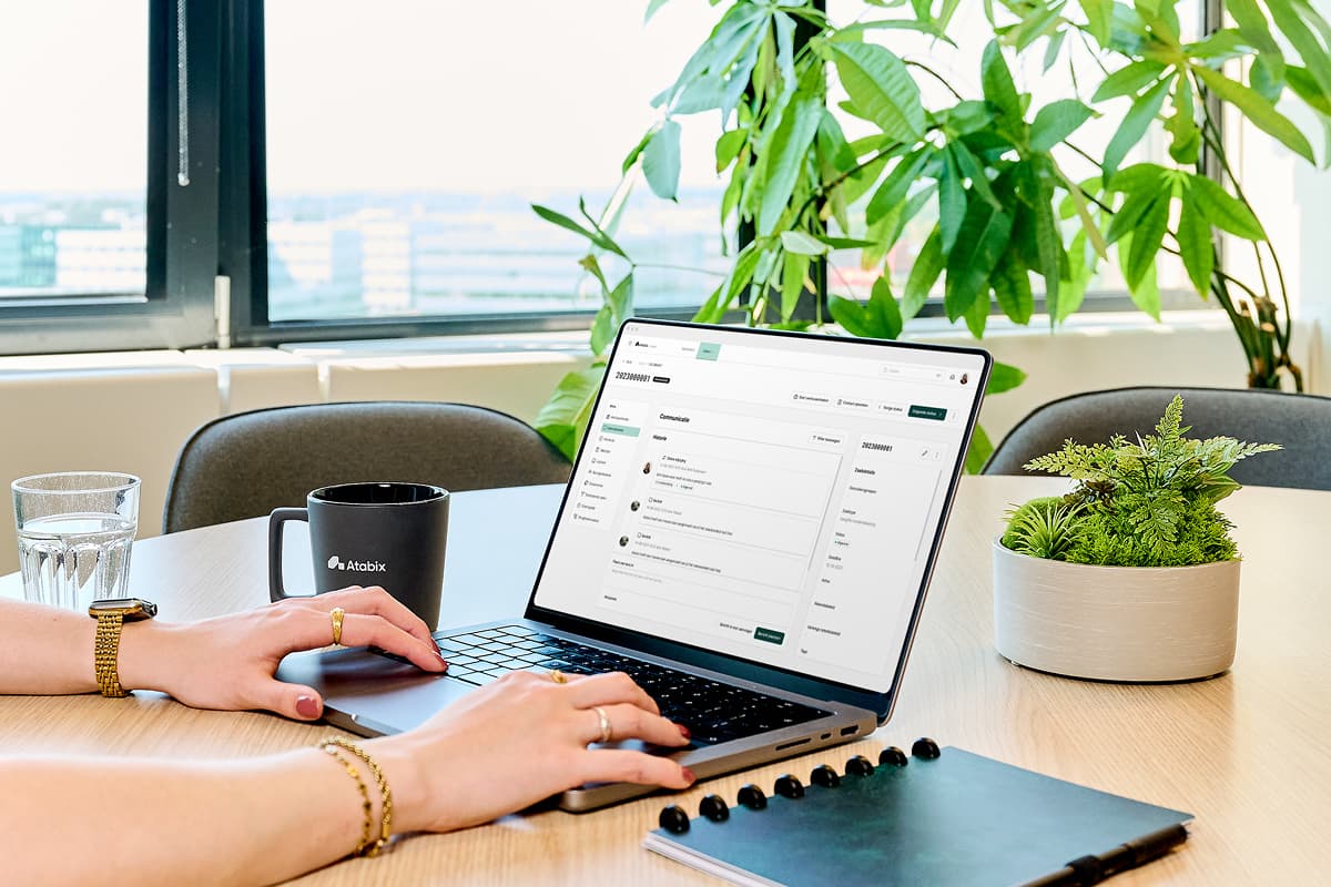 Persoon werkt op een laptop met daarop de Atabix Suite interface. Op het scherm is een communicatieoverzicht zichtbaar binnen een digitaal dossier. Op de vergadertafel staan een Atabix-mok, een glas water, een notitieboek, een pen en een plantenbak. Op de achtergrond grote groene kamerplanten en een raam met uitzicht op kantoorgebouwen.