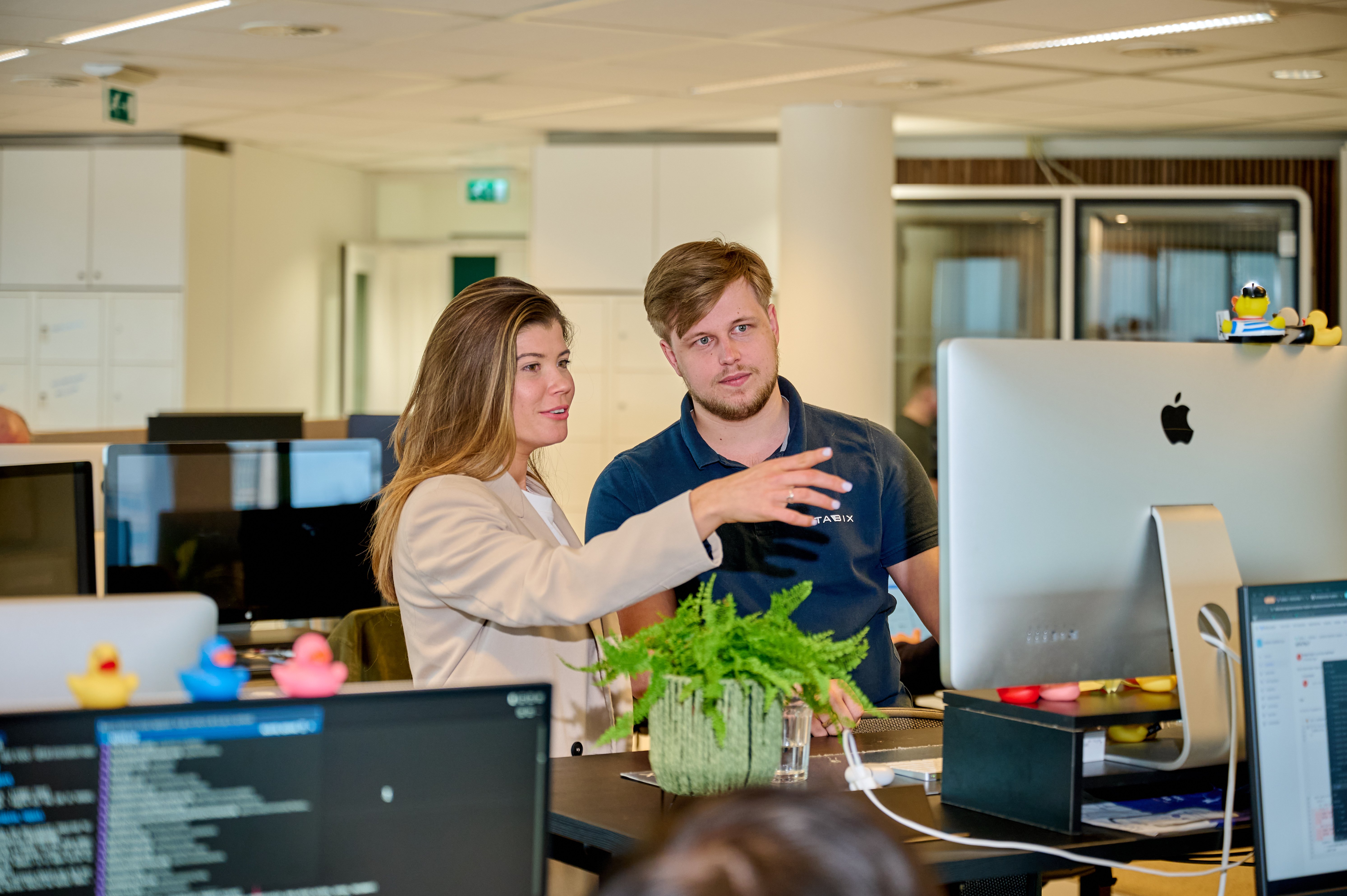 Vrouw en man in kantoorruimte, staan achter een staand bureau en kijken naar een computer, vrouw wijst iets aan op scherm. 